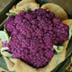 A large head of purple graffiti cauliflower with green leaves, placed on a burlap fabric.
