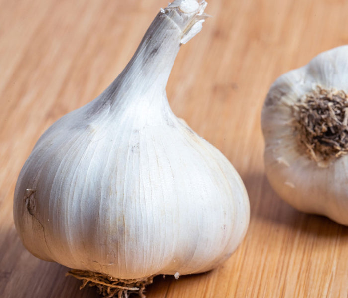 A close-up of a whole blue music garlic bulb on a light wooden surface, with another garlic bulb partially visible in the background.
