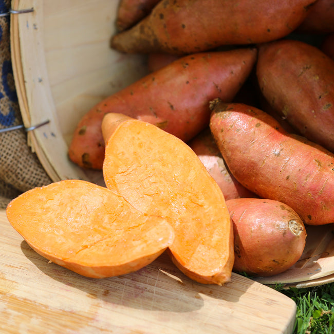 A group of Vardaman sweet potatoes with one cut in half to show the orange flesh, placed on a wooden cutting board with a burlap sack in the background.
