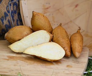 Elongated tubers of white yams with light tan skin and bright white flesh on a wooden cutting board, with a burlap sack in the background.