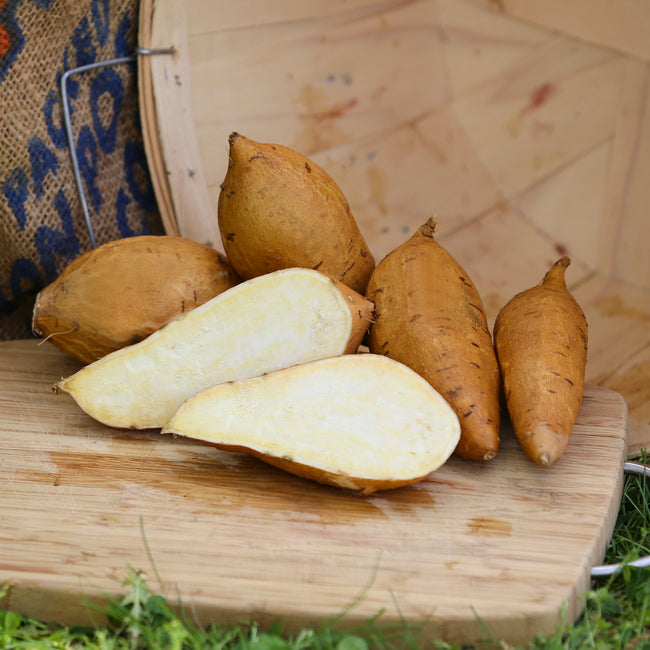 Elongated tubers of white yams with light tan skin and bright white flesh on a wooden cutting board, with a burlap sack in the background.
