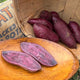 A wooden basket filled with whole purple majesty sweet potatoes, with a few sweet potatoes cut in half and laid on a wooden platter.
